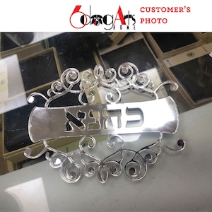 May include: A silver-coloured metal sign with a decorative scroll design and the Hebrew word "ברוכה" in the centre. The sign is on a reflective surface.