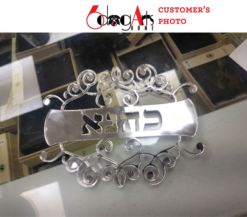 May include: A silver-coloured metal sign with a decorative scroll design and the Hebrew word "ברוכה" in the centre. The sign is on a reflective surface.
