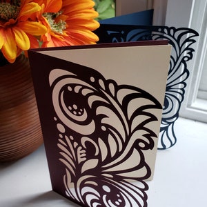 May include: A burgundy card with a white intricate design cut out of the card. The card is standing on a white surface.