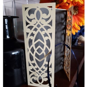 May include: A beige card with a decorative cut-out design. The card is folded in half and has a blue ribbon tied around it. The card is sitting on a wooden surface.