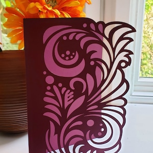 May include: A burgundy card with a white and pink floral design cut out. The design features swirls, leaves, and dots. The card is ready for use with electronic cutting machines. SVG, DXF, EPS, PDR, PNG digital template files.
