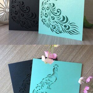 May include: Two peacock-themed greeting cards. One card is black, the other is teal. Each card features a detailed peacock design cut out of the paper. The cards are displayed on a wooden surface. Text on the image reads: SVG, DXF, EPS, PNG CUTTING FILES READY FOR MAJOR ELECTRONIC CUTTING MACHINES.