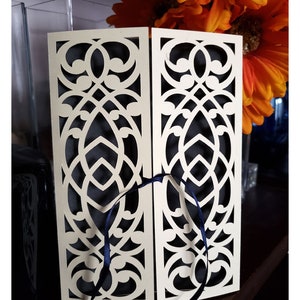 May include: A white cardstock die cut with a decorative, intricate design. The cardstock is folded in half and tied with a blue ribbon. The cardstock is on a wooden surface.