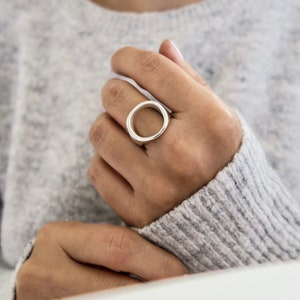 以下が含まれることがあります： 大きな開いた円形のデザインのシルバーリング。指に着用されています。