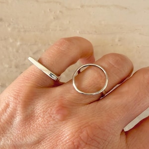 May include: Two silver rings are displayed on a hand. One ring is a simple band with a rectangular top, and the other is a circular ring. The rings are made of a shiny, reflective metal.