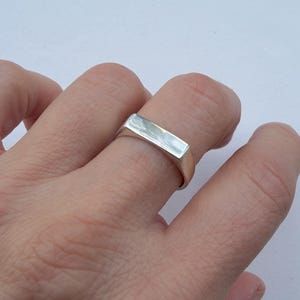 Könnte beinhalten: Ein silberner Ring mit rechteckiger Oberfläche wird an einem Finger getragen. Der Ring hat ein schlichtes, modernes Design und eine polierte Oberfläche. Der Ring ist ein Schmuckstück.