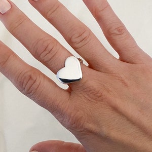 K&ouml;nnte beinhalten: Ein silberner Ring in Herzform an einer Hand mit hellrosa Nagellack.