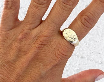 May include: A silver ring featuring a wide, oval-shaped face with a textured, hammered surface. The band is smooth and tapers slightly toward the back, showcasing a metallic finish with subtle indentations across the top surface.