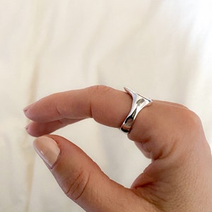 K&ouml;nnte beinhalten: Ein silberner Ring mit einem einzigartigen, abstrakten Design. Der Ring wird an einem Finger getragen und hat eine glatte, polierte Oberfl&auml;che.
