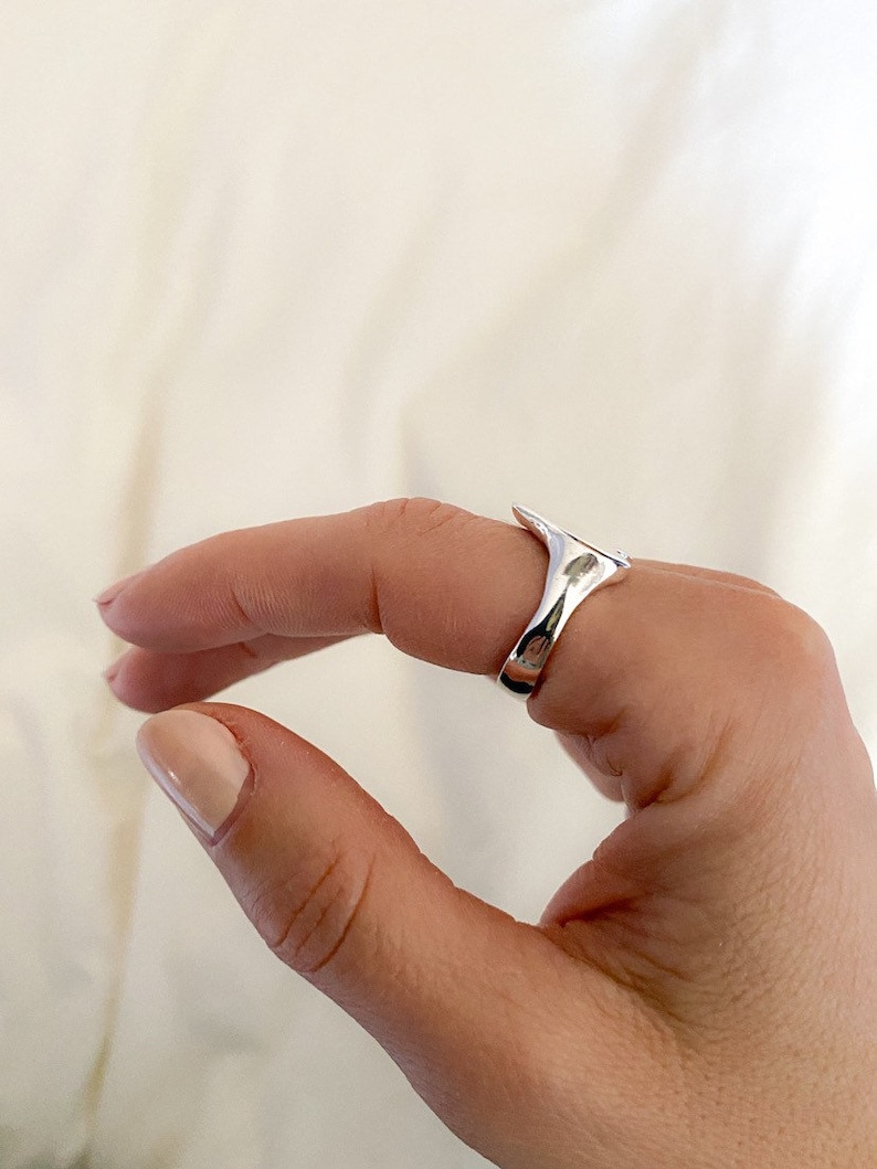 K&ouml;nnte beinhalten: Ein silberner Ring mit einem einzigartigen, abstrakten Design. Der Ring wird an einem Finger getragen und hat eine glatte, polierte Oberfl&auml;che.