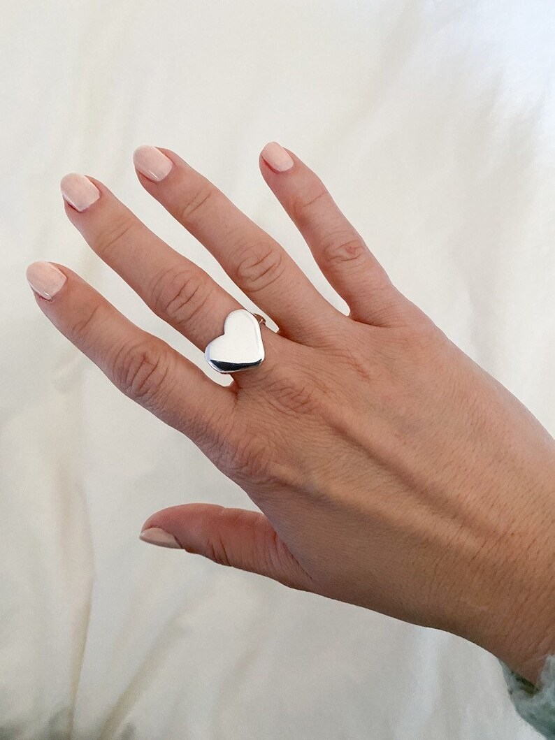 K&ouml;nnte beinhalten: Ein silberner Ring in Herzform an einer Hand mit hellrosa Nagellack.