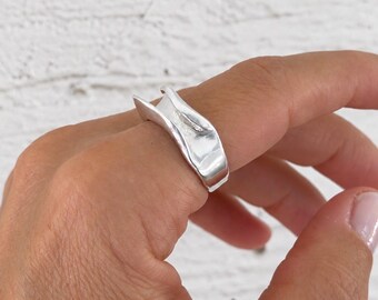 May include: A silver ring with an irregular, wavy, and organic shape. The band features a polished, reflective surface with a distinct curve that creates a gap at the top, showcasing a smooth, metallic finish that wraps around the finger.