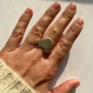 K&ouml;nnte beinhalten: Ein silberner Ring in Herzform an einem Finger. Der Ring ist gro&szlig; und hat eine glatte, polierte Oberfl&auml;che.