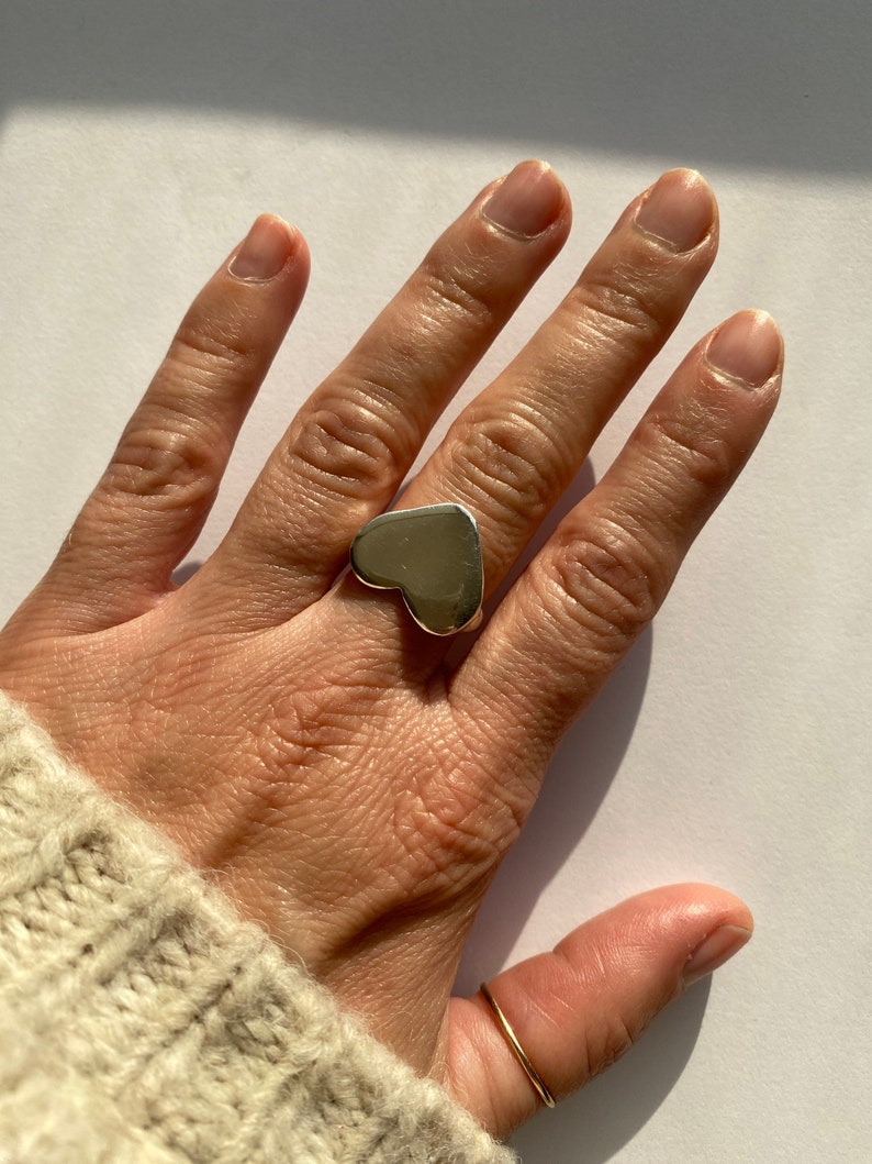 K&ouml;nnte beinhalten: Ein silberner Ring in Herzform an einem Finger. Der Ring ist gro&szlig; und hat eine glatte, polierte Oberfl&auml;che.