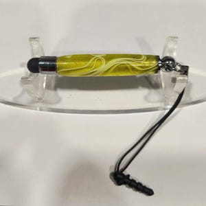 May include: A yellow and white swirl stylus pen with a black tip and a silver metal band. The pen has a black cord with a plug at the end. The stylus is displayed on a clear, oval-shaped stand.