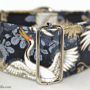 Op de afbeelding: Een brede hondenhalsband met een zwarte achtergrond met een wit kraanvogelontwerp met gouden en grijze bloemaccenten. De halsband heeft een zilveren metalen gesp en hardware. De website galgocotilla.com staat onderaan afgedrukt.
