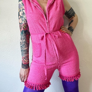 May include: A pink terrycloth jumpsuit with a zipper closure, a tie at the waist, and fringe trim around the neckline, sleeves, and hem.
