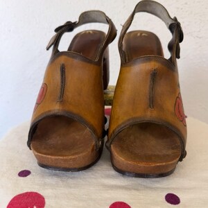 May include: A pair of vintage brown leather clogs with a floral design. The open-toe sandals have a wooden sole and a strap that buckles around the ankle. The heels are approximately 3 inches high.