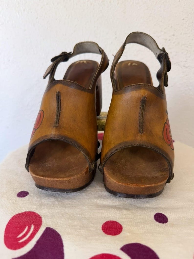 May include: A pair of vintage brown leather clogs with a floral design. The open-toe sandals have a wooden sole and a strap that buckles around the ankle. The heels are approximately 3 inches high.