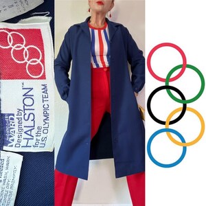 以下が含まれることがあります： 「Designed by HALSTON™ for the U.S. OLYMPIC TEAM」と記されたラベルが付いたネイビーブルーのロングコート。コートは赤と白のストライプのシャツと赤いパンツの上に着用されています。背景にはオリンピックの輪があります。