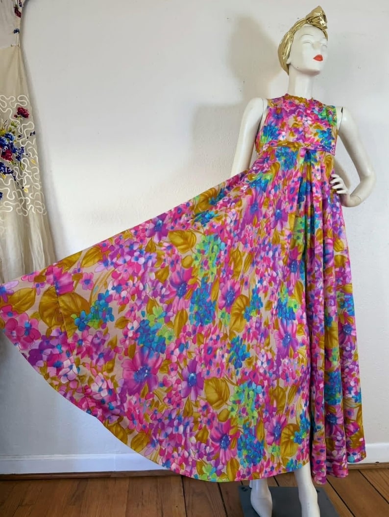 May include: A vintage floral maxi dress in shades of pink, purple, blue, and yellow. The sleeveless dress features a high neckline and a flowing skirt. The dress is displayed on a mannequin with a gold head wrap.