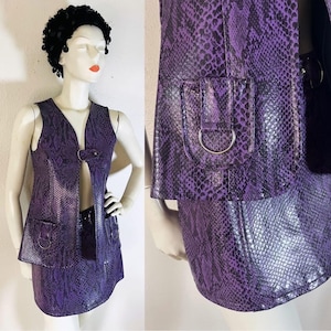 Puede incluir: Un conjunto de dos piezas con estampado de piel de serpiente púrpura. El conjunto incluye un chaleco con cierre metálico y una minifalda a juego. El chaleco tiene dos bolsillos con aros metálicos. El conjunto se exhibe en un maniquí.