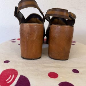 May include: A pair of vintage brown leather sandals with thick, wooden block heels. The sandals feature ankle straps with buckles and a wide strap across the top of the foot. The heels are approximately 3 inches (7.6 cm) tall.