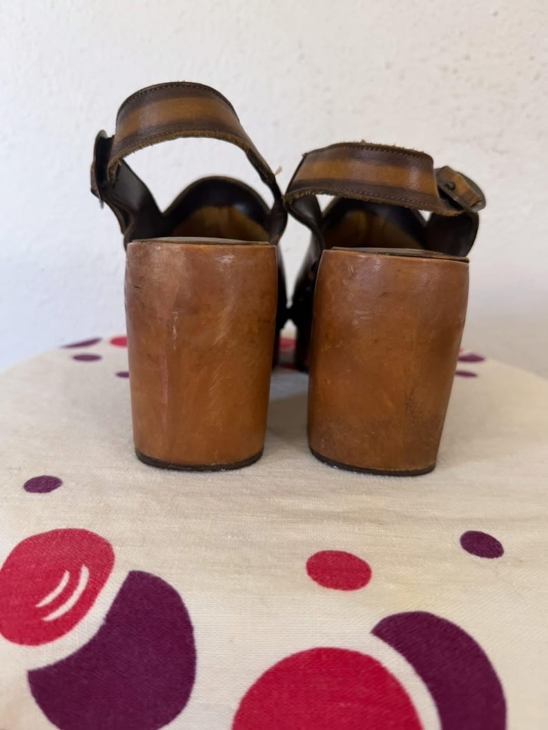 May include: A pair of vintage brown leather sandals with thick, wooden block heels. The sandals feature ankle straps with buckles and a wide strap across the top of the foot. The heels are approximately 3 inches (7.6 cm) tall.
