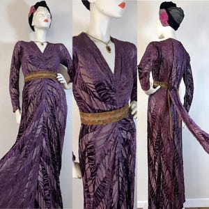 May include: A vintage purple velvet gown with a V-neck and long sleeves. The dress features a leaf-like pattern and is cinched at the waist with a gold-colored belt. A matching fabric sash drapes from the back. The dress is displayed on a mannequin.