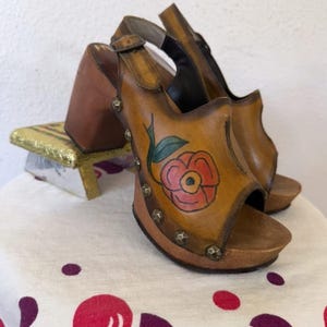 May include: A pair of vintage-style open-toe sandals with a wooden platform and heel. The tan leather uppers feature a hand-painted red flower design with green leaves and are adorned with silver studs. The sandals have a buckle closure.