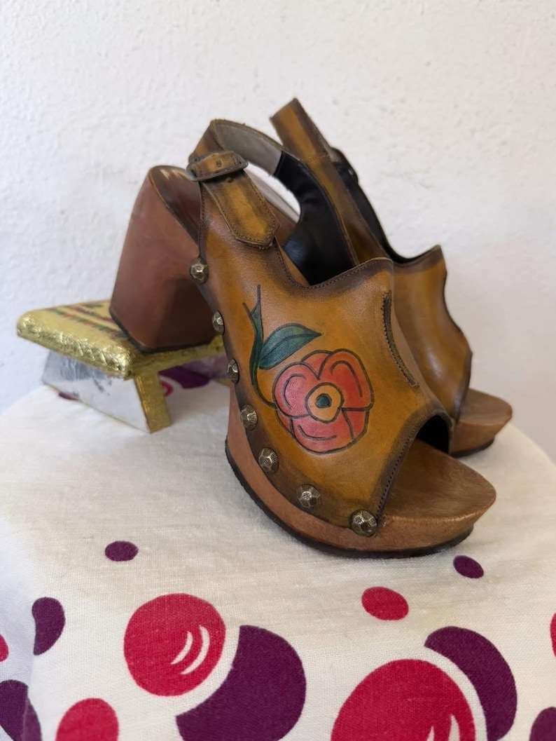 May include: A pair of vintage-style open-toe sandals with a wooden platform and heel. The tan leather uppers feature a hand-painted red flower design with green leaves and are adorned with silver studs. The sandals have a buckle closure.