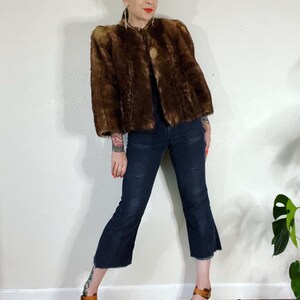 May include: A brown faux fur jacket with a cropped length and a stand-up collar. The jacket has a slightly puffed shoulder design and is worn with dark blue jeans and brown sandals.