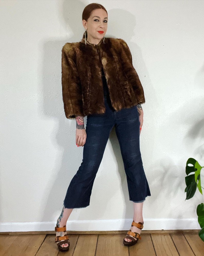 May include: A brown faux fur jacket with a cropped length and a stand-up collar. The jacket has a slightly puffed shoulder design and is worn with dark blue jeans and brown sandals.