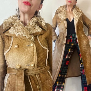 Puede incluir: Un abrigo largo de gamuza marrón con cuello de piel de oveja y cinturón. El abrigo tiene cierre de doble botonadura y dos bolsillos en la parte delantera.