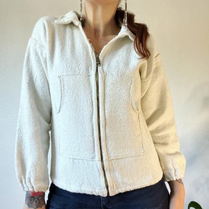 May include: A white zip-up jacket with a collared neckline and a single pocket on the front. The jacket has a textured, slightly fuzzy fabric.