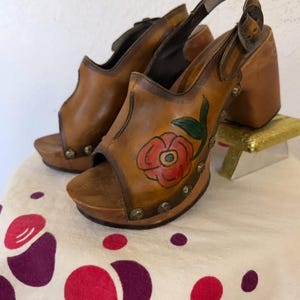 May include: A pair of vintage brown leather clogs with a floral design. The open-toe shoes have a wooden platform and heel, with a strap around the ankle. The floral design features a red rose with green leaves. The shoes are decorated with small metal studs.
