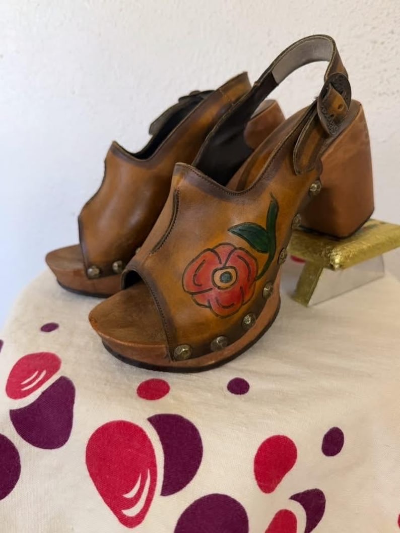 May include: A pair of vintage brown leather clogs with a floral design. The open-toe shoes have a wooden platform and heel, with a strap around the ankle. The floral design features a red rose with green leaves. The shoes are decorated with small metal studs.
