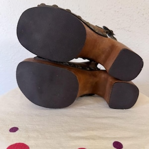 May include: Pair of brown wooden platform sandals with dark rubber soles. The sandals feature a buckle closure and decorative metal studs. The wooden heels are approximately 4 inches high.
