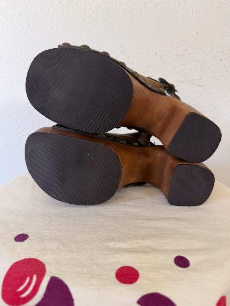 May include: Pair of brown wooden platform sandals with dark rubber soles. The sandals feature a buckle closure and decorative metal studs. The wooden heels are approximately 4 inches high.