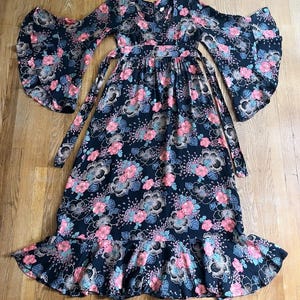 May include: A long black maxi dress with a floral print in shades of pink, blue, and green. The dress has a V-neckline, long sleeves with wide bell cuffs, and a tie at the waist. The skirt has a ruffled hem.