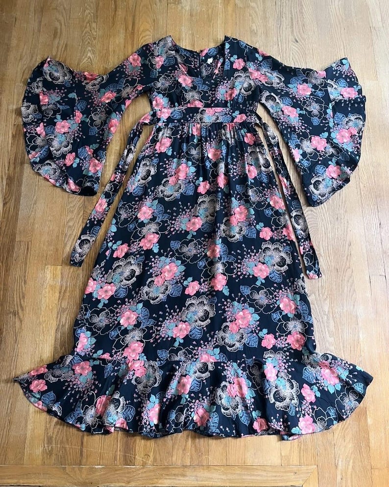 May include: A long black maxi dress with a floral print in shades of pink, blue, and green. The dress has a V-neckline, long sleeves with wide bell cuffs, and a tie at the waist. The skirt has a ruffled hem.