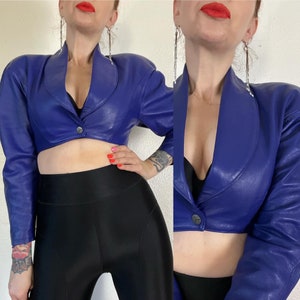 May include: A cropped blue leather jacket with a pointed collar and long sleeves. The jacket has a button closure and is worn over a black top and black pants.