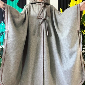 Op de afbeelding: Een grijze capuchon cape met bruine stiksels en een strik sluiting. De cape heeft een lang, vloeiend ontwerp en is gemaakt van een zachte, warme stof.