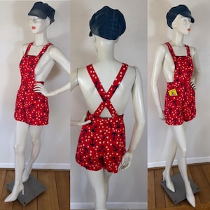 Könnte beinhalten: Ein roter Jumpsuit mit einem sich wiederholenden Muster aus dunkelblauen Ankern und weißen Sternen. Das Kleidungsstück hat einen eckigen Ausschnitt, breite Schulterträger, die sich am Rücken kreuzen, und kurze Hosenbeine. Ein kleines gelbes Etikett ist an der Seitennaht befestigt.