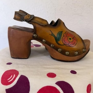 May include: Vintage brown leather clogs with a floral design. The open-toe sandals feature a high wooden heel, a buckle strap, and decorative metal studs. The painted flower is red with green leaves.