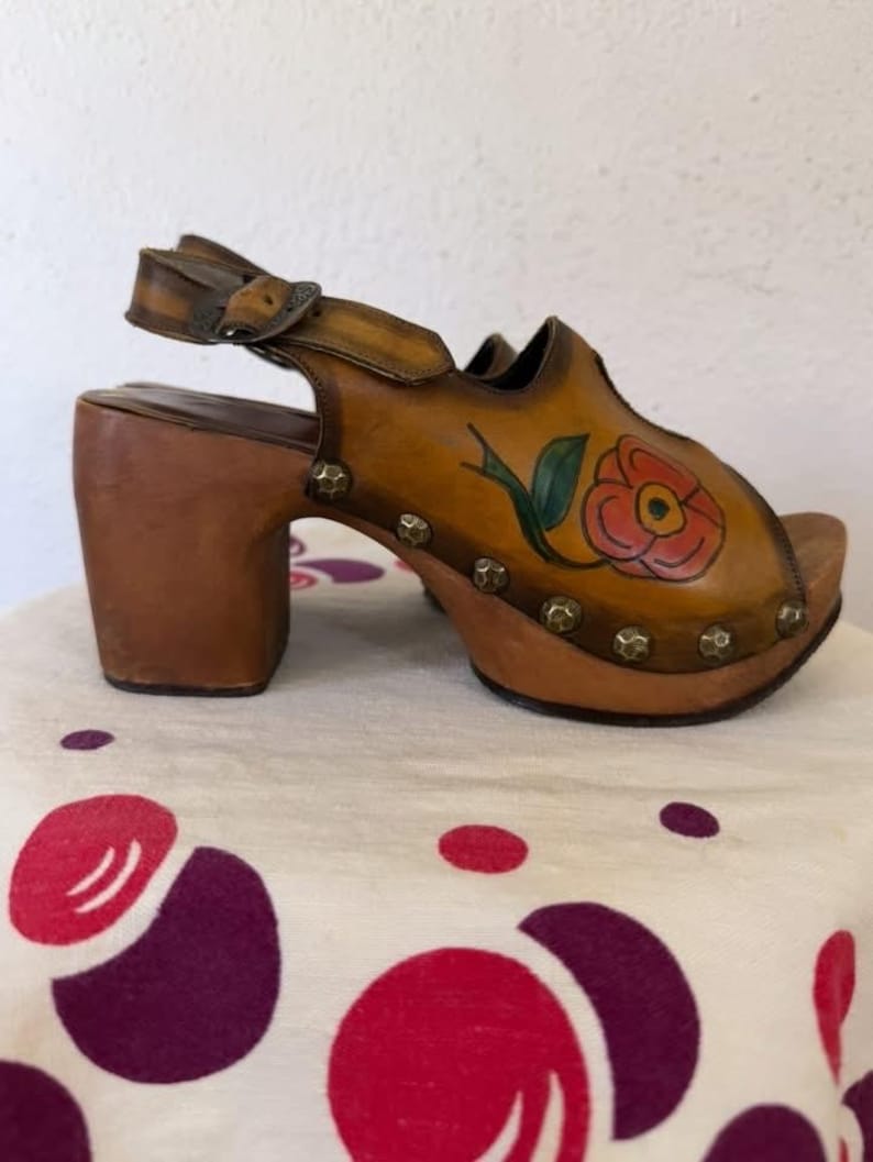 May include: Vintage brown leather clogs with a floral design. The open-toe sandals feature a high wooden heel, a buckle strap, and decorative metal studs. The painted flower is red with green leaves.
