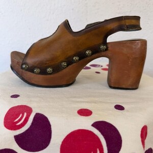 May include: A brown leather clog sandal with a wooden platform and heel. The sandal features a peep-toe design, a strap around the ankle, and decorative metal studs. The heel measures approximately 3 inches.
