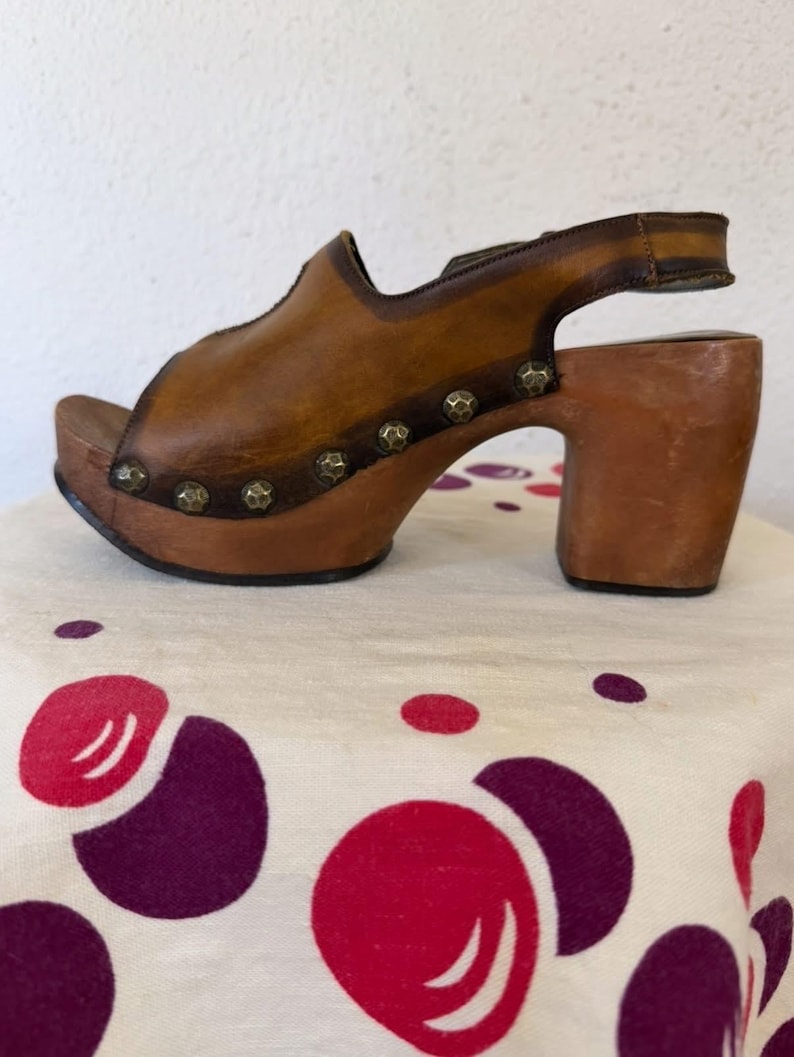 May include: A brown leather clog sandal with a wooden platform and heel. The sandal features a peep-toe design, a strap around the ankle, and decorative metal studs. The heel measures approximately 3 inches.
