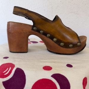 May include: A vintage brown leather clog sandal with a wooden platform and heel. The sandal features a peep-toe design, a slingback strap, and decorative metal studs. The heel measures approximately 3 inches.