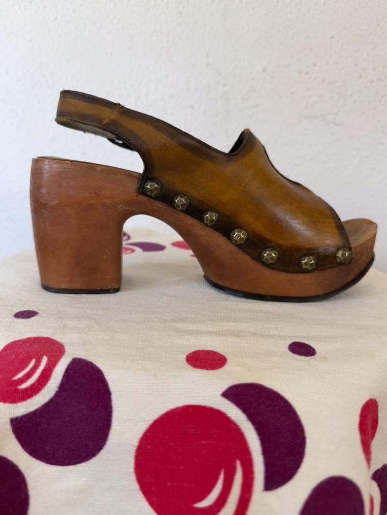May include: A vintage brown leather clog sandal with a wooden platform and heel. The sandal features a peep-toe design, a slingback strap, and decorative metal studs. The heel measures approximately 3 inches.